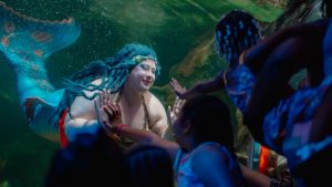 A performer dressed as a mermaid with a shimmering blue tail and matching hair interacts playfully with children through the glass at an aquarium, pressing hands together in a magical undersea moment